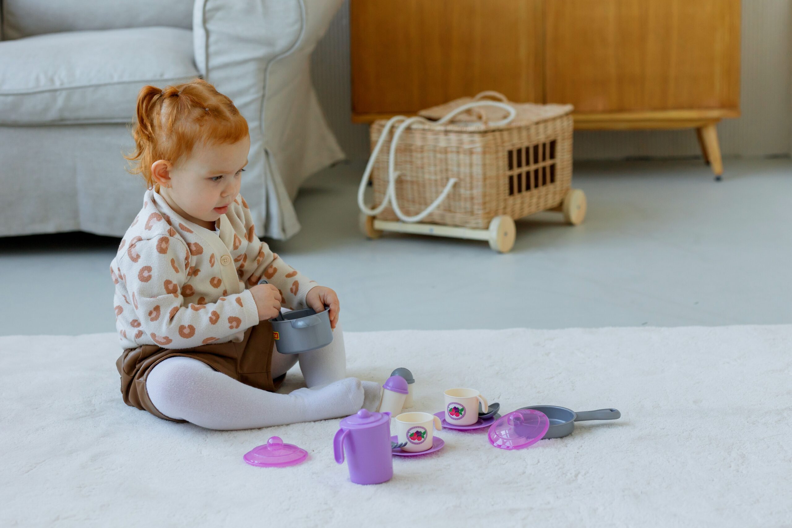 Criança pequena sentada em tapete branco felpudo na sala, brincando com panelinhas e xícaras de brinquedo lilás